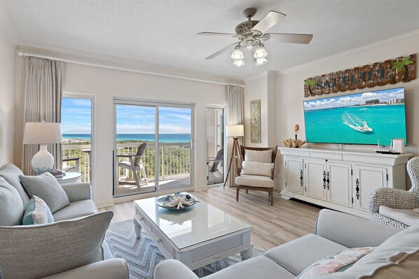 A spacious living room with a large TV, comfortable seating, a ceiling fan, and a view of the ocean through the sliding doors.