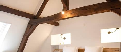 Ein Schlafzimmer mit schrägen Wänden, Holzbalken, einem Bett mit weißer Bettwäsche, Nachttischlampen und einem Schreibtisch mit Stuhl.