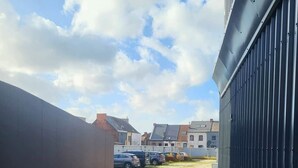 Uma passagem pavimentada leva a uma área de estacionamento com carros, um edifício com fachada escura e casas residenciais ao fundo.