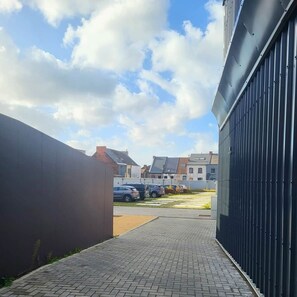 A paved pathway leading to a parking area with cars, a building with a dark facade, and residential houses in the background. - Gite à binche - A l'Orange Sanguine (BINCHE)