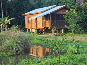 A wooden cabin with a metal roof, situated by a pond surrounded by lush greenery. - Freshwater Creek Cabanas (Hopkins)