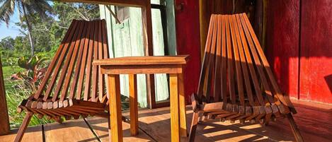 Two wooden lounge chairs and a small table on a wooden deck with a red wall in the background.