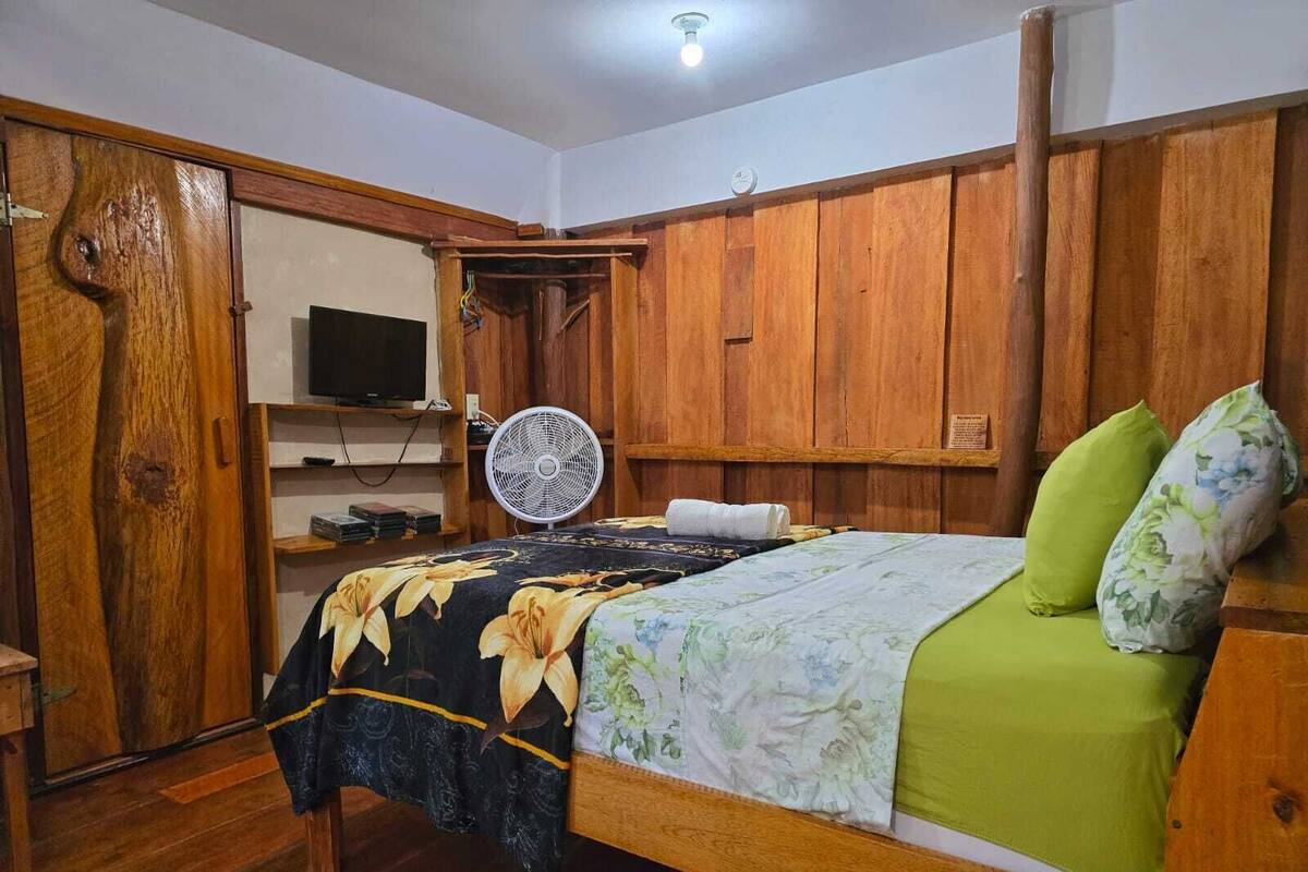 A room with a wooden bed, a fan, and a television on a shelf.