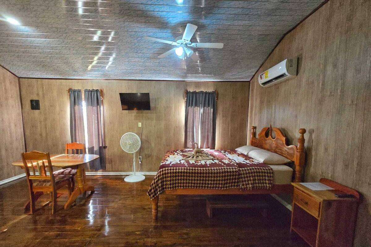 A room with a wooden floor, a bed with a patterned bedspread, a wooden chair, a small table, a ceiling fan, and an air conditioning unit.
