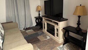 A living room with a beige sofa, a flat-screen TV on a wooden stand, two lamps, and a fireplace. - Charming 1-bedroom apartment with AC, WiFi in beautiful Fairport (Fairport)