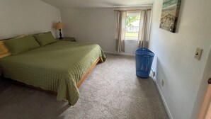 A bedroom with a bed, a nightstand, a lamp, a window with curtains, a blue laundry basket, and a painting on the wall. - Charming 1-bedroom apartment with AC, WiFi in beautiful Fairport (Fairport)