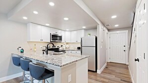 A modern kitchen with white cabinets, a granite countertop, and a built-in microwave. There are two bar stools and a refrigerator. - Cozy Boise Stay: Central Spot (Boise)