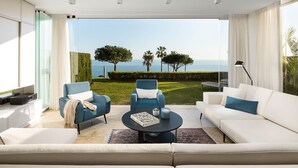 Una sala de estar moderna con un sofá blanco, dos sillones azules y una mesita de centro. Un ventanal ofrece vistas al mar y a un jardín cuidado.