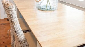 A dining area with a wooden table, wicker chairs, a glass vase with a plant, and a life ring on the wall. - CB Surf House|Charming Beach House 2mins to Beach (Carolina Beach)