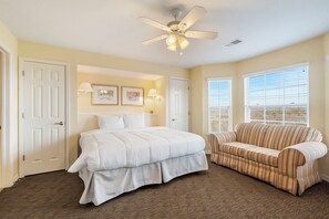 A bedroom with a bed, a sofa, a ceiling fan, and two windows.
