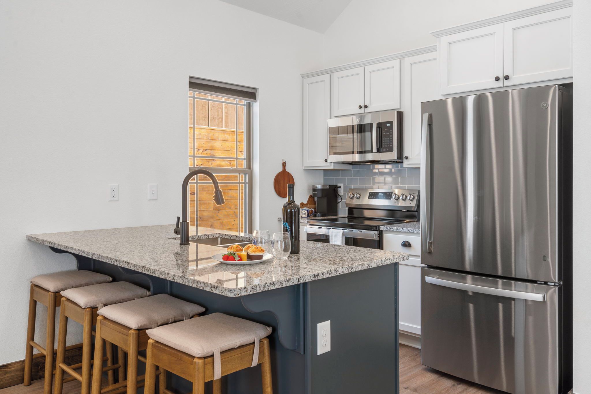 A modern kitchen with a stainless steel refrigerator, a microwave, and a stove. There are bar stools and a window with blinds.