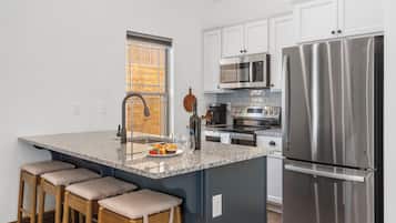 A modern kitchen with a stainless steel refrigerator, a microwave, and a stove. There are bar stools and a window with blinds.