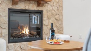 A stone wall with a fireplace, a round wooden table with a plate of food, and a bottle of wine.