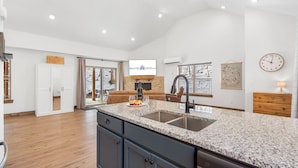 A spacious kitchen with granite countertops, a built-in sink, and a large island. There is a dining area with a brown sofa and a TV mounted on the wall.