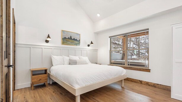 A bedroom with a large bed, wooden flooring, a window with a view of trees, and a painting on the wall.