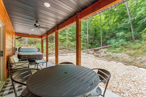 Uma área externa coberta com mesa e cadeiras, uma banheira de hidromassagem e vista para uma área arborizada.