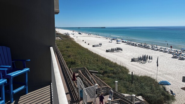 A beach view from a balcony with blue chairs, a wooden deck, and a pier in the distance.