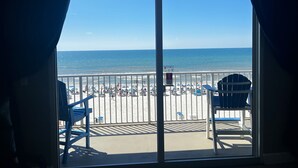 A balcony with a view of the beach, featuring a railing, a ceiling fan, and two blue chairs.