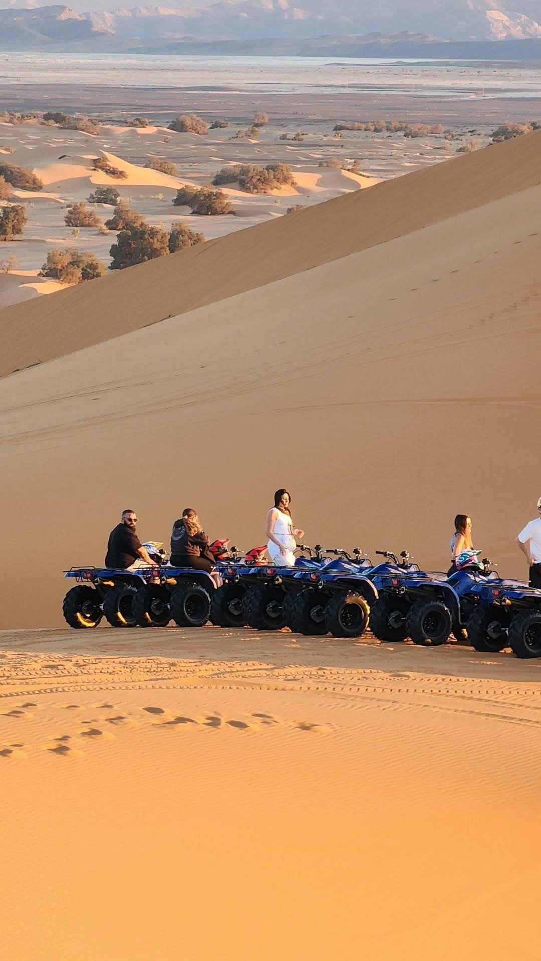 A group of people on ATVs on a sand dune with a vast desert landscape in the background.