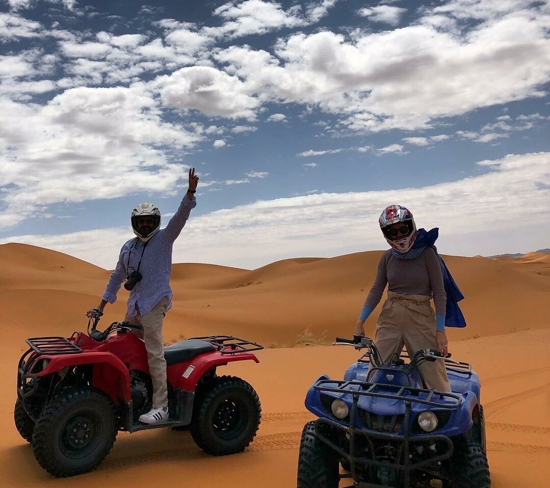 Two people on ATVs in a desert with sand dunes and a partly cloudy sky.
