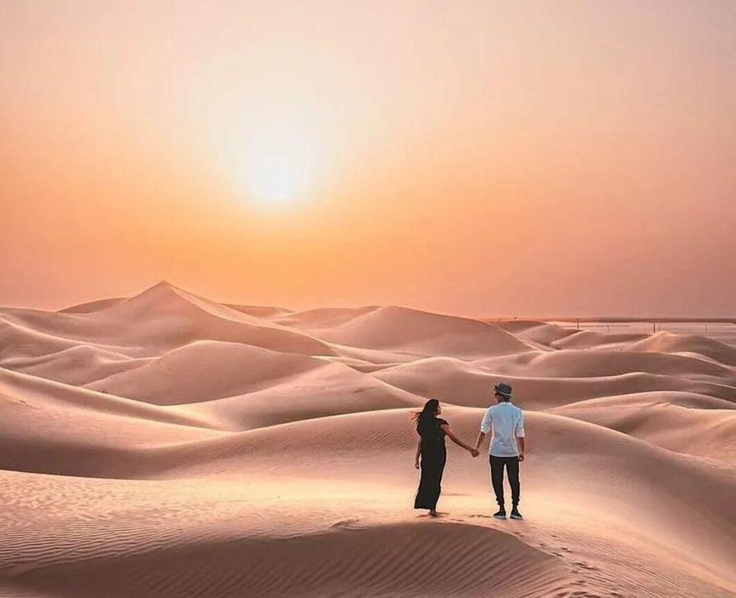 Un couple se tient sur une dune de sable, avec un coucher de soleil en arrière-plan.