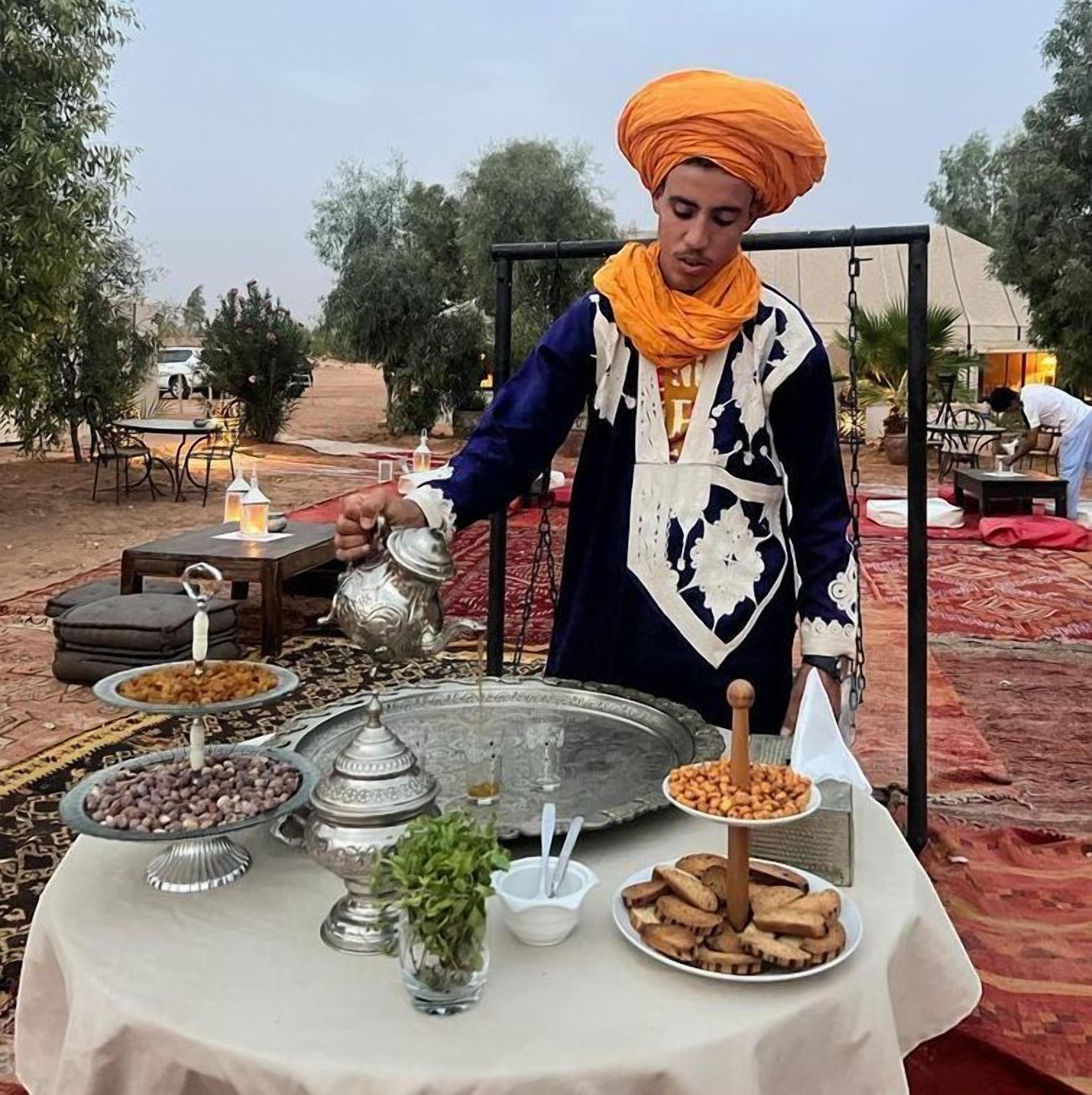 Una persona sirve comida en una mesa al aire libre, con diversos platos y una carpa al fondo.