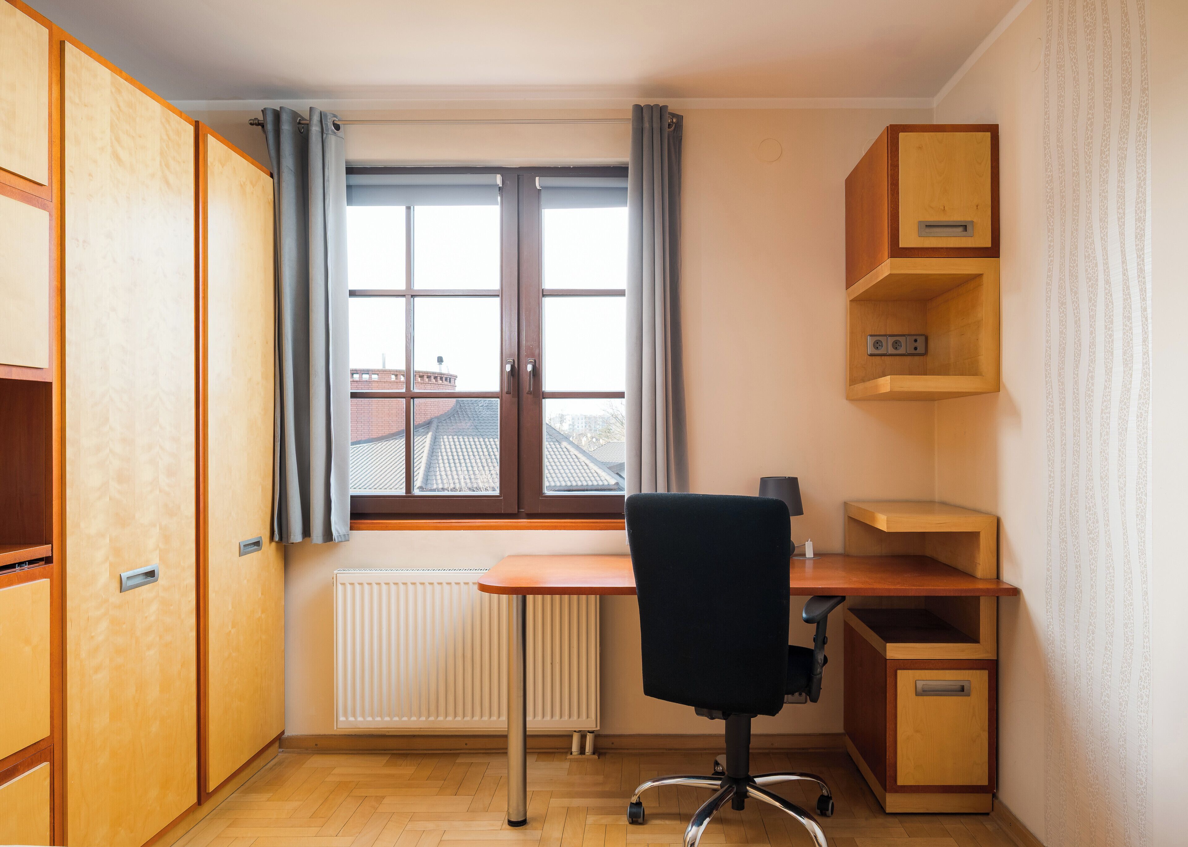 Een kleine, goed verlichte kamer met een houten bureau, een zwarte bureaustoel, een radiator en een raam met gordijnen.