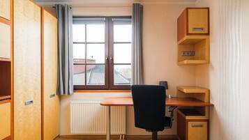 Een kleine, goed verlichte kamer met een houten bureau, een zwarte bureaustoel, een radiator en een raam met gordijnen.