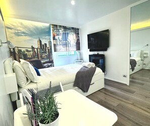 Una camera d'albergo moderna con un letto, una TV a schermo piatto, un'ampia finestra con vista sulla città, un tavolino con una pianta e uno specchio a parete.
