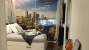 A bedroom with a cityscape mural, a bed with storage drawers, a chair, and a small table.