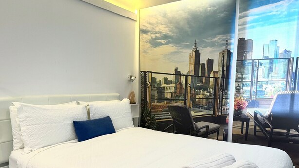 A hotel room with a large bed, a cityscape mural on the wall, a balcony with a chair, and a table with a vase of flowers.