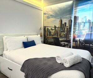 A hotel room with a large bed, a cityscape mural on the wall, a balcony with a chair, and a table with a vase of flowers.