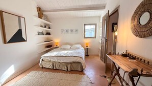 A bedroom with a bed, bedside lamps, a wooden dresser, and a mirror. There are shelves with various items and a window providing natural light.