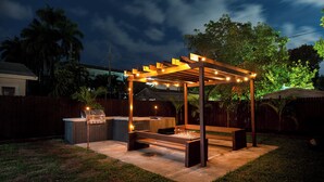A covered outdoor patio with a grill and seating area, surrounded by a wooden fence and trees.