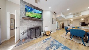 A modern living room with a fireplace, a TV mounted on a stone wall, and a dining area with blue chairs. - Ski-in Ski-out  | Updated | High Ceilings | Gold Creek 204 (Angel Fire)