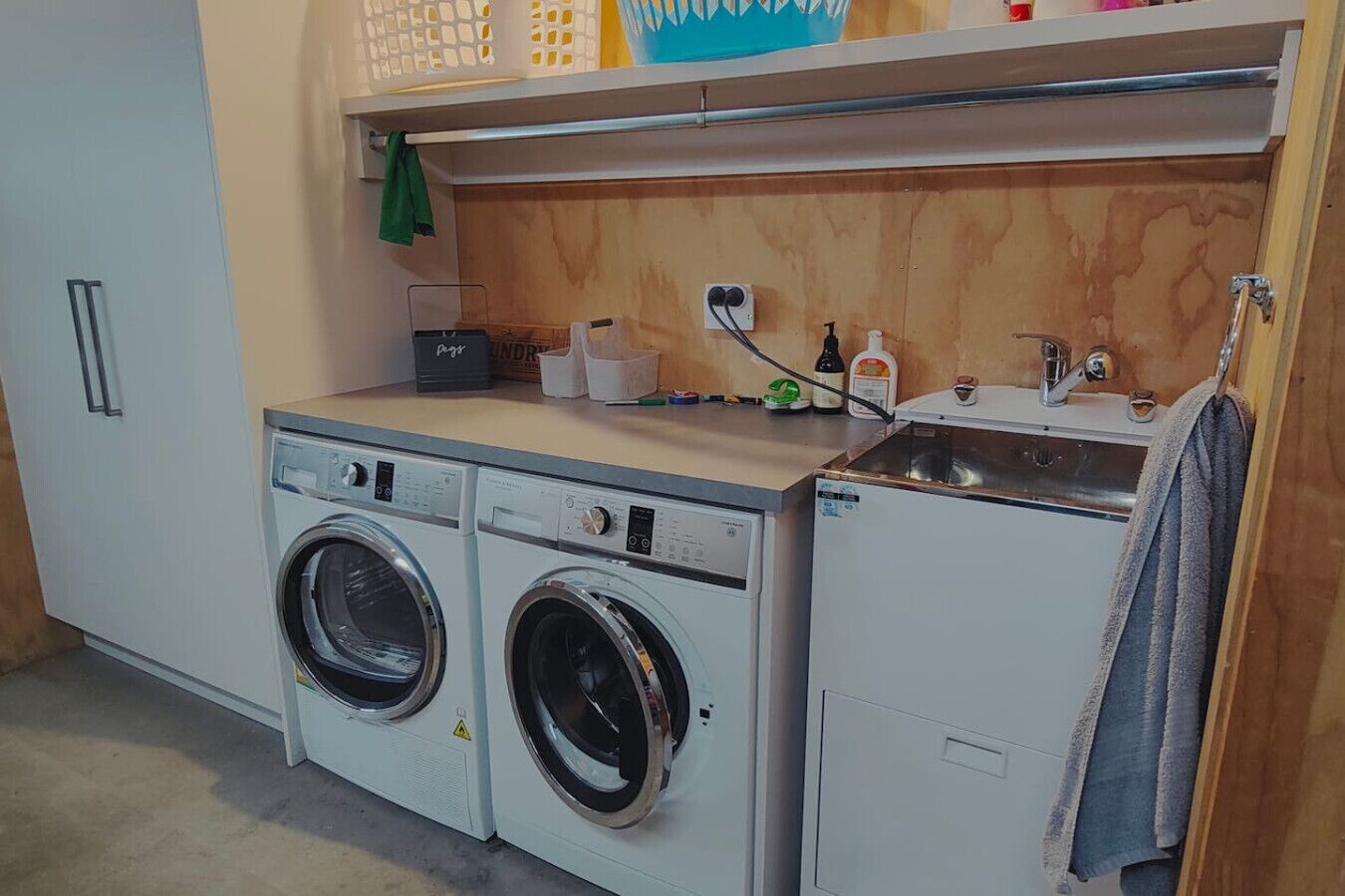 A laundry area with two front-loading washing machines, a sink, and a shelf above with various items.