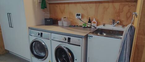 A laundry area with two front-loading washing machines, a sink, and a shelf above with various items.