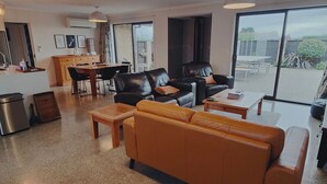 Un vaste espace de vie comprenant un canapé, une table basse et des fauteuils, un coin repas avec une table et des chaises, et une vue sur l’extérieur grâce à de grandes fenêtres.
