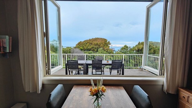 A dining area with a table and chairs, a vase with flowers, and a view of an outdoor deck with a railing and chairs. - Riverton Beachhouse View (Riverton / Aparima)