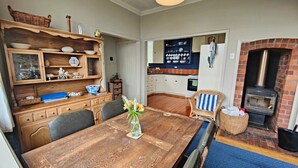 A rustic kitchen with a wooden table, shelves with dishes, a blue chair, and a brick fireplace. - Riverton Beachhouse View (Riverton / Aparima)