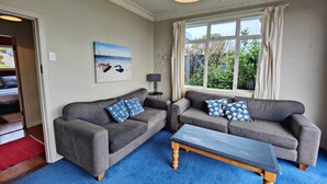 A living room with two grey sofas, a blue carpet, a coffee table, a painting on the wall, and a window with curtains. - Riverton Beachhouse View (Riverton / Aparima)