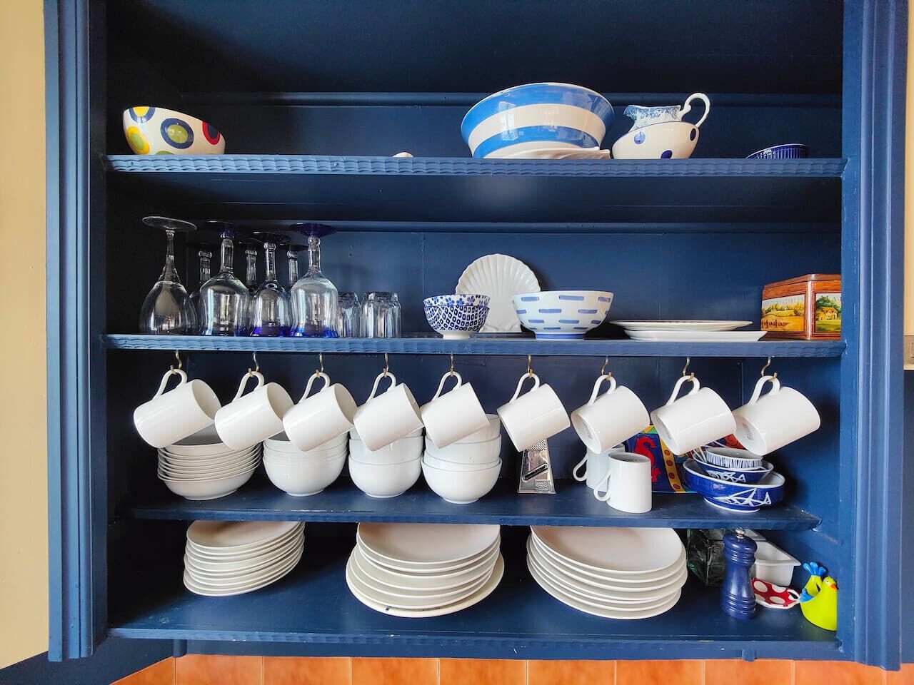 Uma estante azul com pratos, copos e um pequeno brinquedo, tudo em tons de branco e azul.
