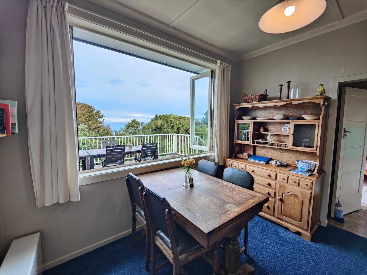 Een kamer met een eettafel en stoelen, een houten kast met planken, een raam met uitzicht op het landschap, en een plafondlamp.