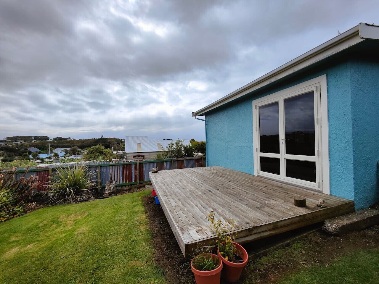 Ein blaues Haus mit einer Holzterrasse, einem großen Fenster und einem umzäunten Garten.