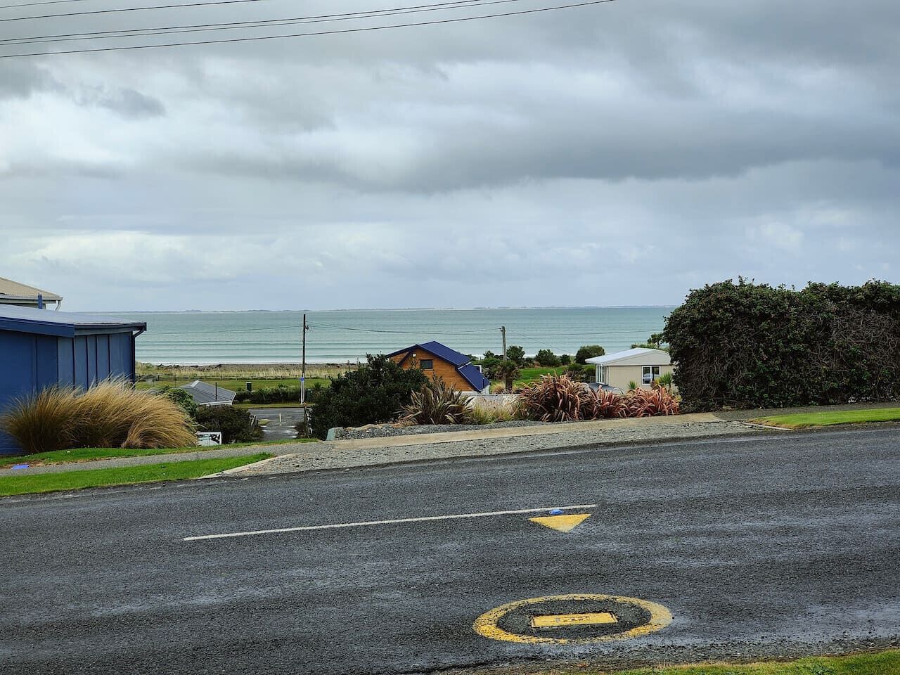 A coastal road with a blue building on the left, a house with a blue roof in the center, and a view of the sea in the background.