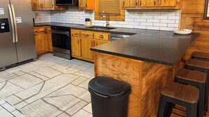 A kitchen with wooden cabinets, a stainless steel refrigerator, a microwave, and a sink. There are stools at the island and a trash can. - Dockside Cabin | RRG | Hot Tub | Kayaks | Fire Pit (Campton)
