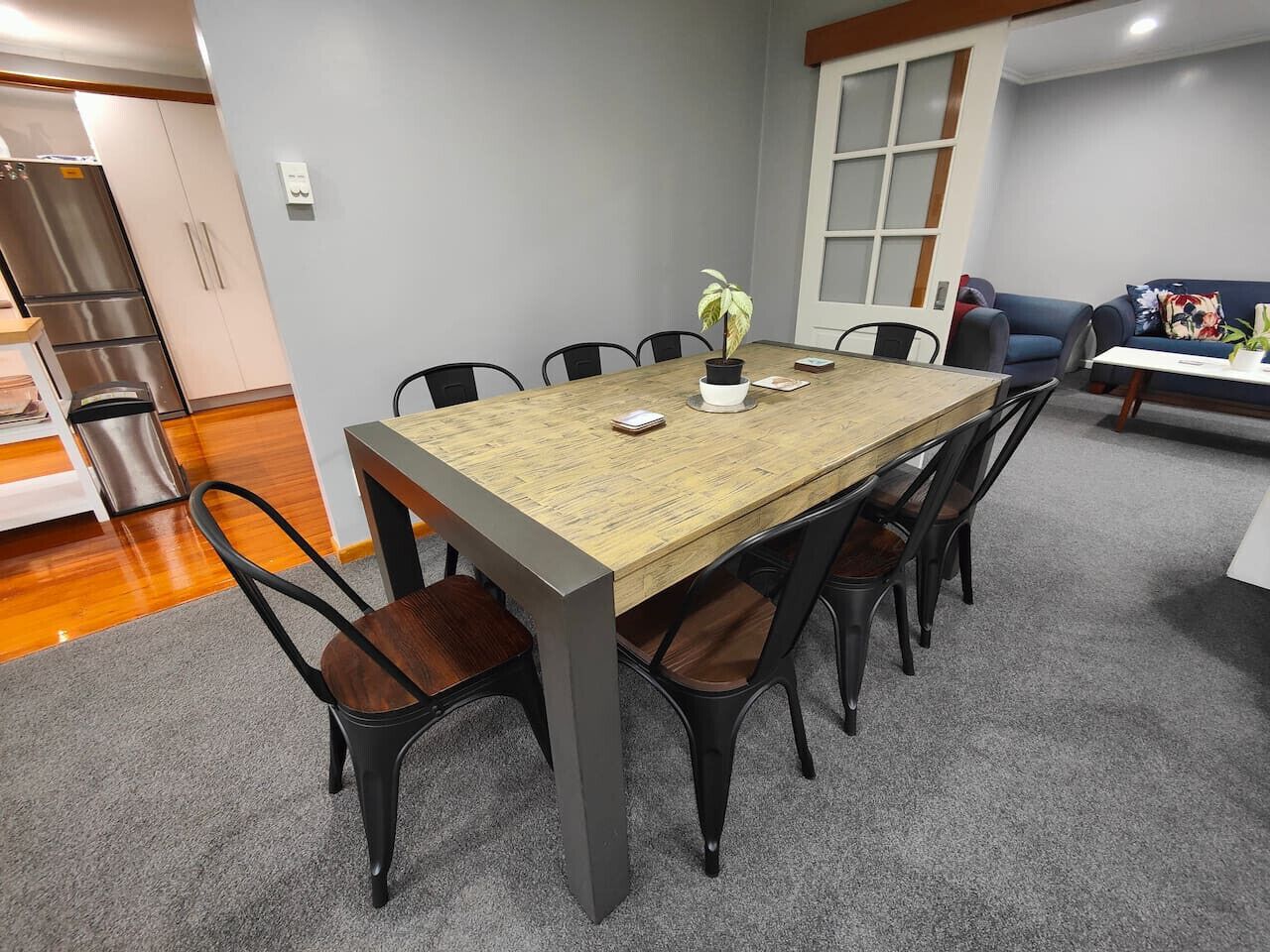 A dining area with a rectangular table, six chairs, a potted plant, and a sofa in the background.