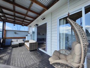 A covered outdoor area with a wooden deck, wicker furniture, and a hanging chair. - Rusty’s Riverton Retreat (Riverton / Aparima)