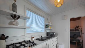 Eine kleine Küche mit weißen Schränken, einem Gasherd, einer Spüle und einem Toaster. Ein Fenster mit Jalousien, eine Hängeleuchte und eine kleine Ablagefläche mit Küchenutensilien sind ebenfalls vorhanden.