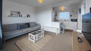 Ein kompakter Wohnraum mit grauem Sofa, einem weißen Couchtisch, einer Küchenzeile mit weißen Schränken und einem Esstisch.